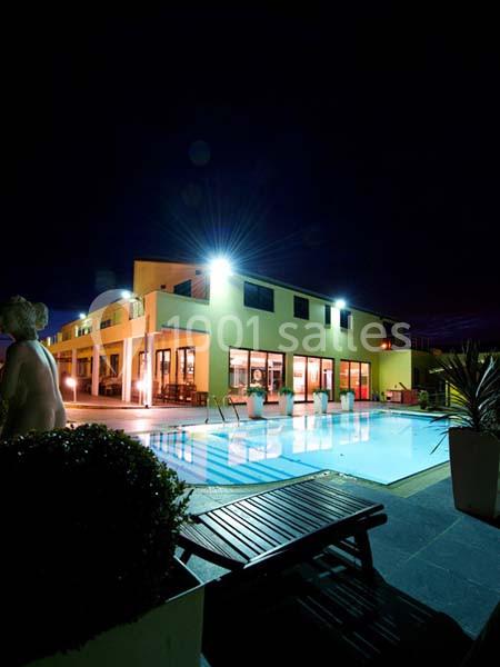 Vue nocturne d'une piscine éclairée devant un bâtiment moderne avec des baies vitrées et une terrasse aménagée.