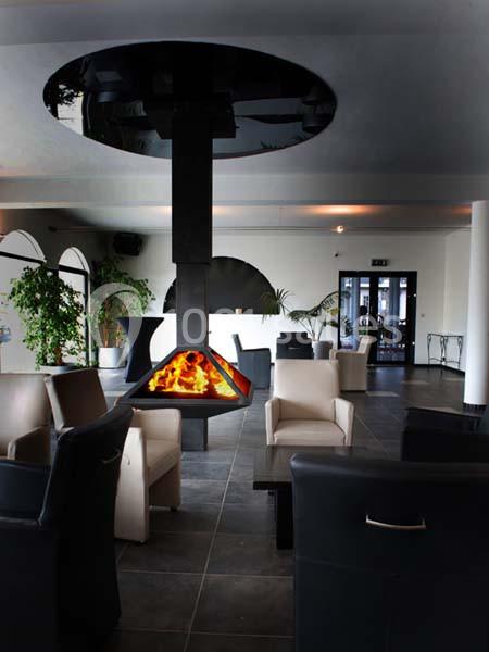 Cheminée suspendue moderne avec flammes vives au centre d'un salon élégant, entourée de fauteuils et de plantes.