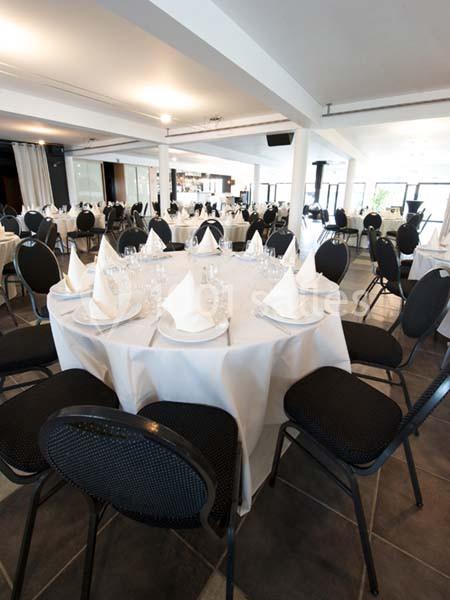 Salle de réception lumineuse avec des tables rondes dressées de nappes blanches, serviettes pliées et chaises noires.