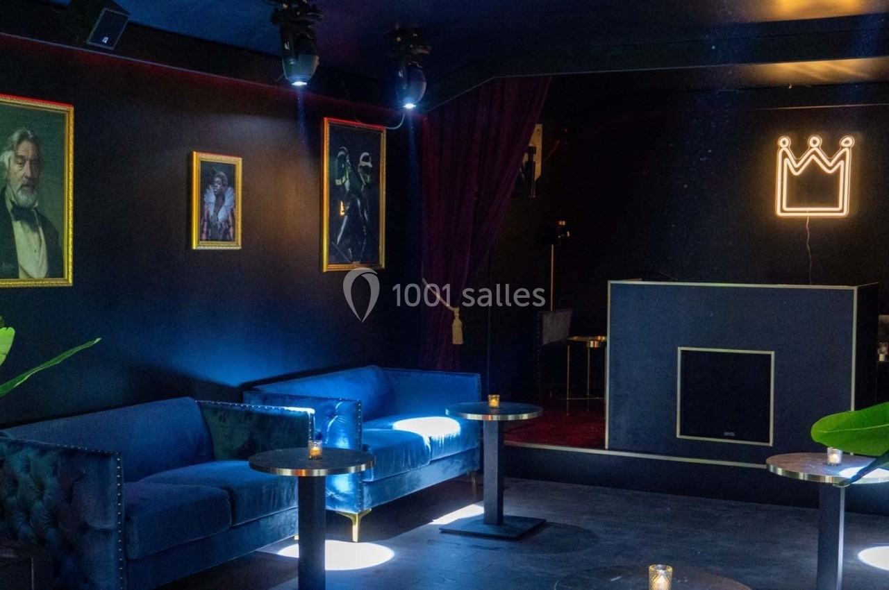 Salle sombre avec canapés bleus, tables basses, plantes, tableaux encadrés et enseigne lumineuse en forme de couronne.