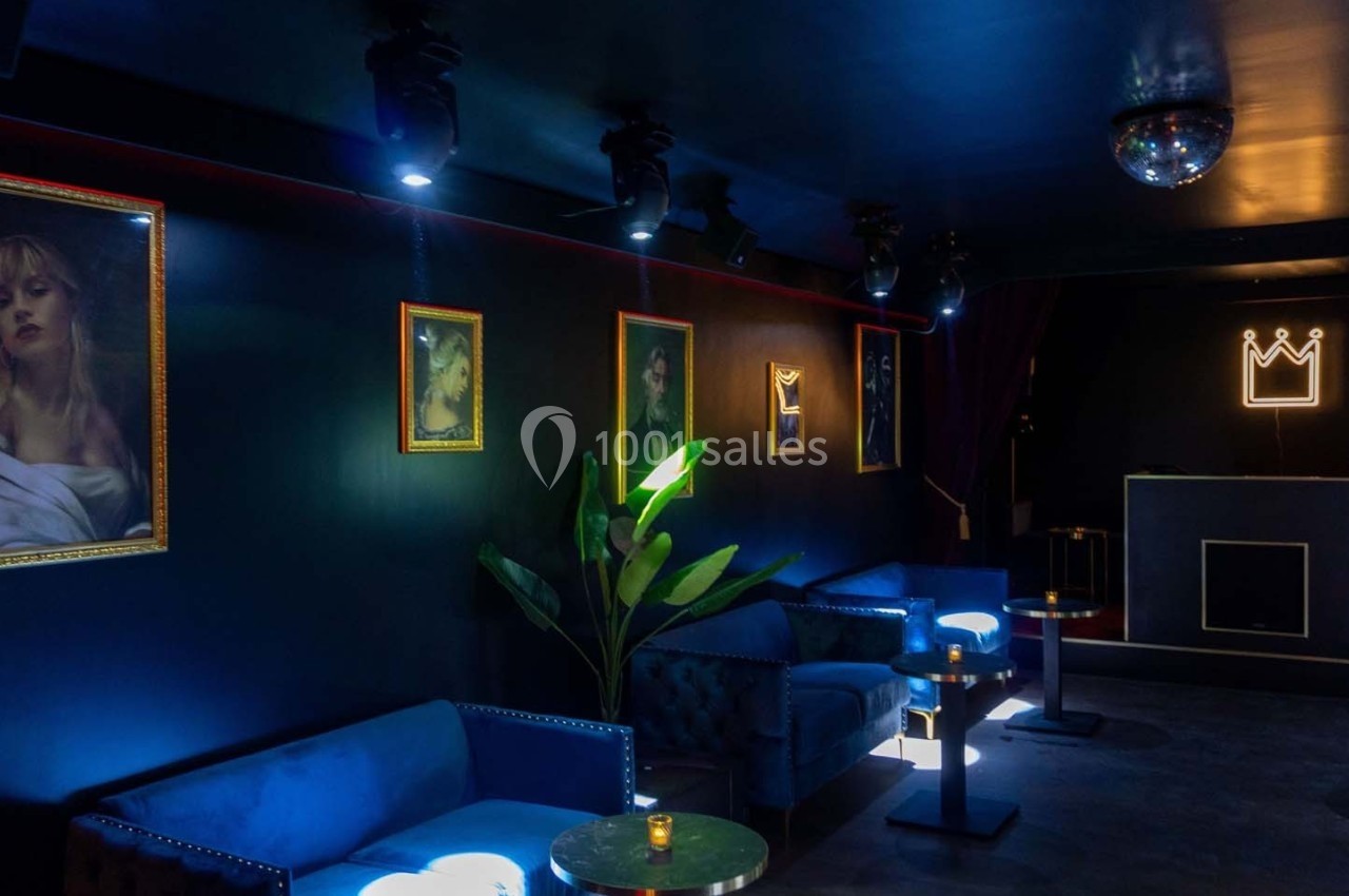 Salle sombre avec canapés bleus, tables rondes, portraits aux murs, plante verte et éclairage tamisé.