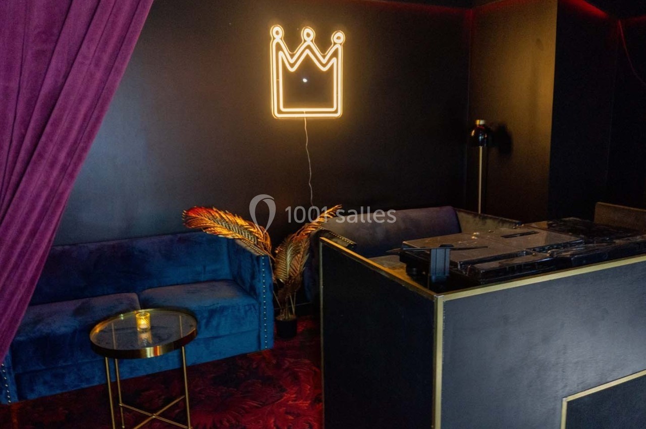 Espace lounge sombre avec canapé bleu, table basse, décoration dorée et néon en forme de couronne sur le mur.