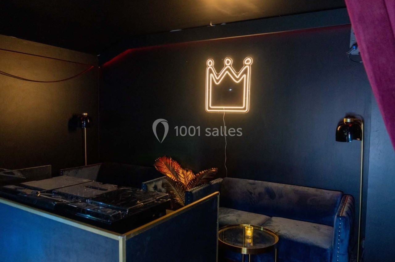 Espace lounge sombre avec canapé bleu, table basse, décoration lumineuse en forme de couronne et rideau rouge.