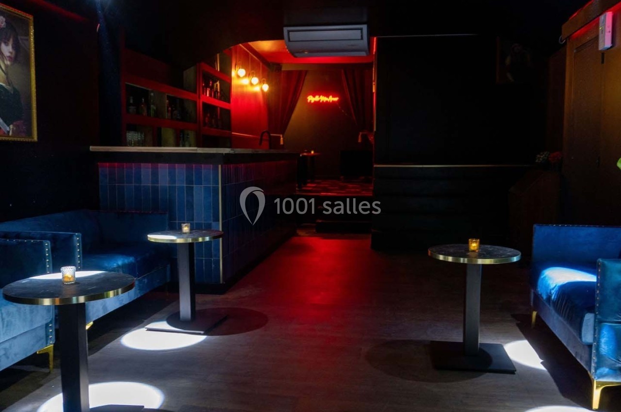 Salle sombre avec canapés bleus, tables basses, éclairage tamisé et néon rouge au fond.