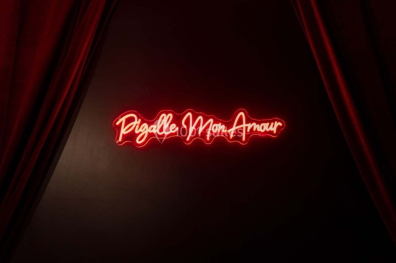 Enseigne néon rouge ’Pigalle Mon Amour’ sur un mur sombre encadrée par des rideaux rouges.