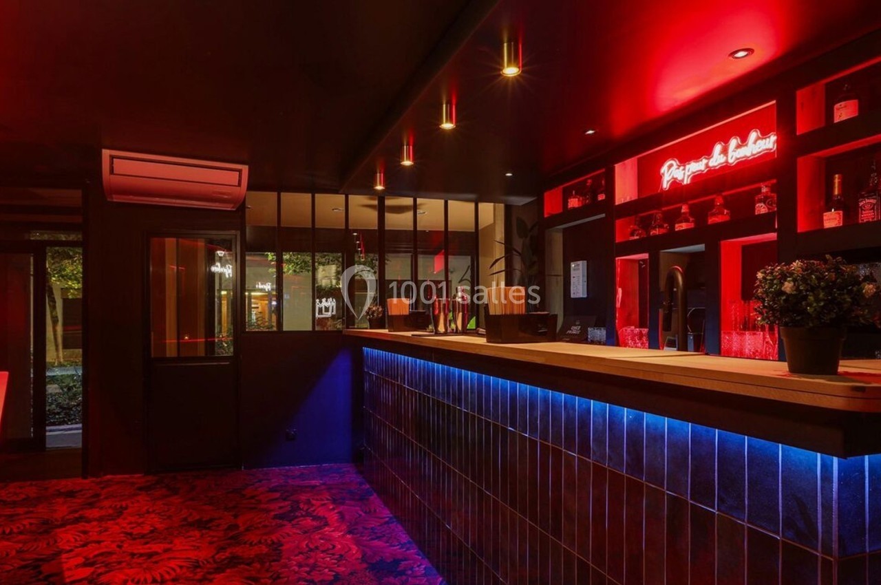 Bar moderne avec éclairage rouge et bleu, comptoir en bois, étagères à bouteilles et décoration végétale.