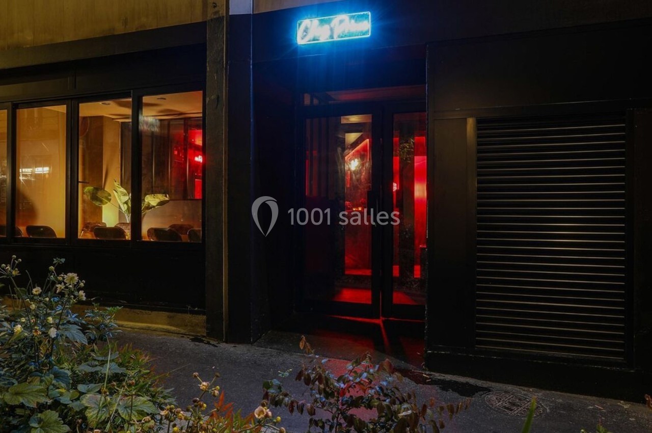 Façade d'un établissement avec une entrée vitrée, éclairée par une lumière rouge et une enseigne lumineuse bleue.