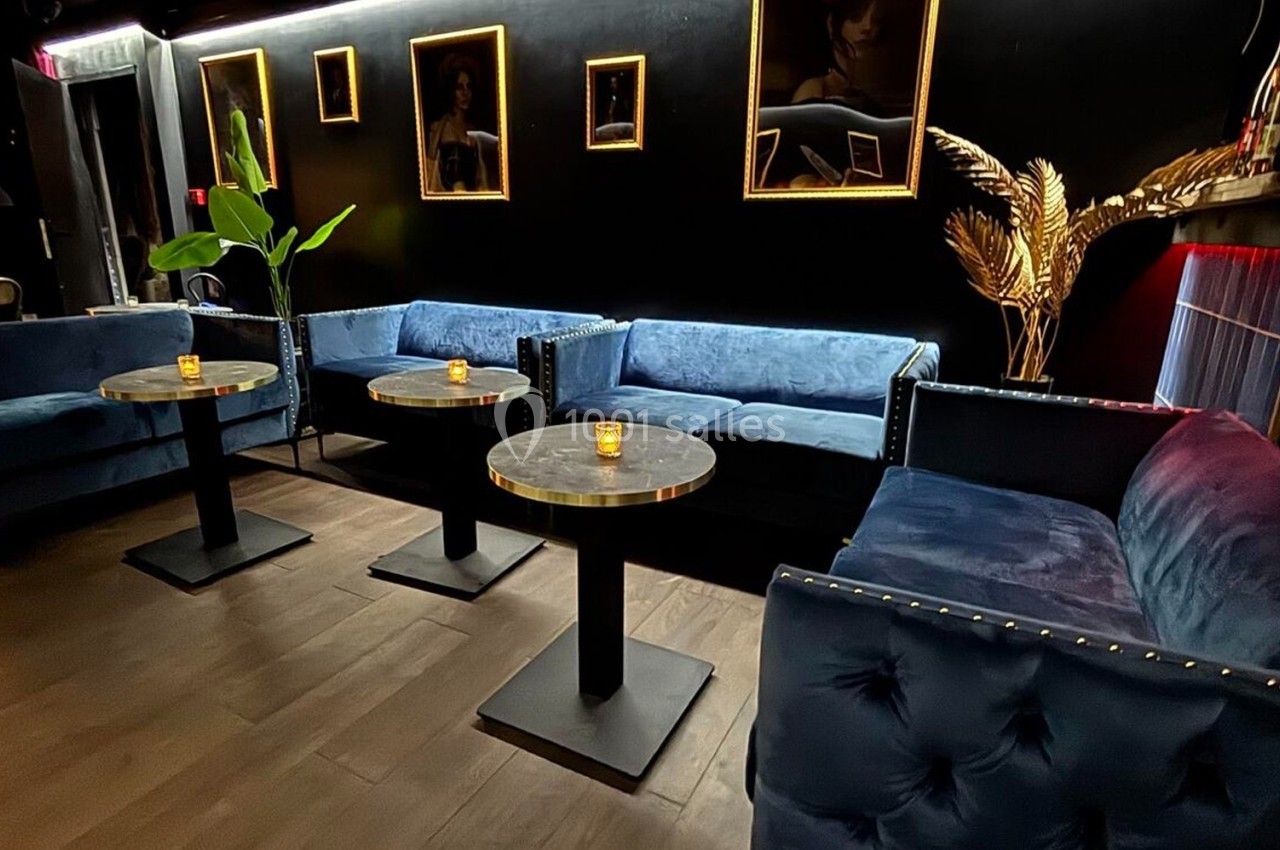 Canapés bleus en velours, tables rondes avec bougies, murs noirs décorés de cadres dorés et éclairage tamisé.