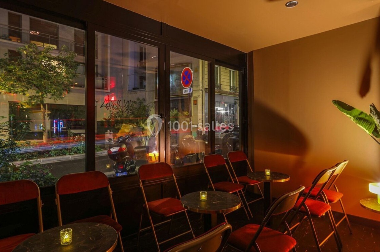 Salle de café intimiste avec chaises pliantes, petites tables rondes et vue sur une rue éclairée la nuit.