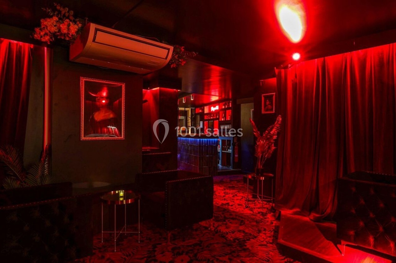 Salle sombre avec éclairage rouge, rideaux épais, fauteuils noirs et un bar illuminé en arrière-plan.
