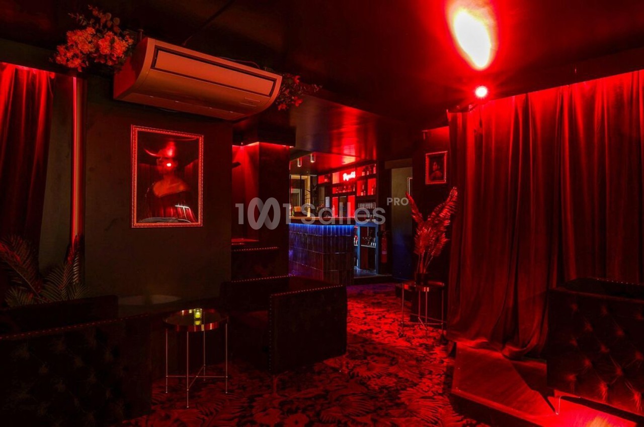 Salle sombre avec éclairage rouge, rideaux épais, fauteuils noirs et un bar illuminé en arrière-plan.