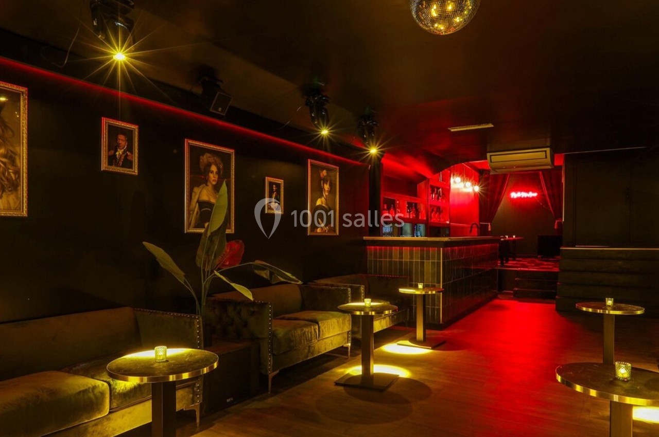 Salle de bar intimiste avec éclairage tamisé rouge, canapés, tables basses et portraits encadrés sur les murs.
