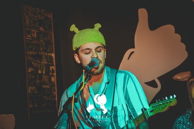 Miniature Charlys Un chanteur interprète sur scène, micro à la bouche, éclairé par une lumière chaude, avec un public en arrière-plan.