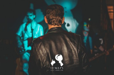 Miniature Charlys Un chanteur interprète sur scène, micro à la bouche, éclairé par une lumière chaude, avec un public en arrière-plan.