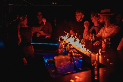 Miniature Cocktail Club Deux femmes chantent et dansent dans une ambiance festive, entourées d'autres personnes dans un lieu éclairé.