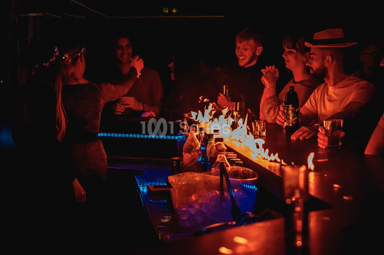 Des personnes discutent et boivent autour d'un bar illuminé par des flammes dans une ambiance sombre.