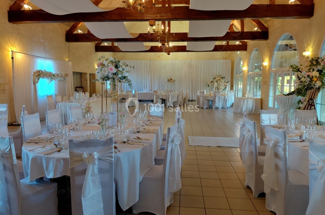 Salle de réception décorée pour un mariage, avec tables rondes dressées, nappes blanches et arrangements floraux.