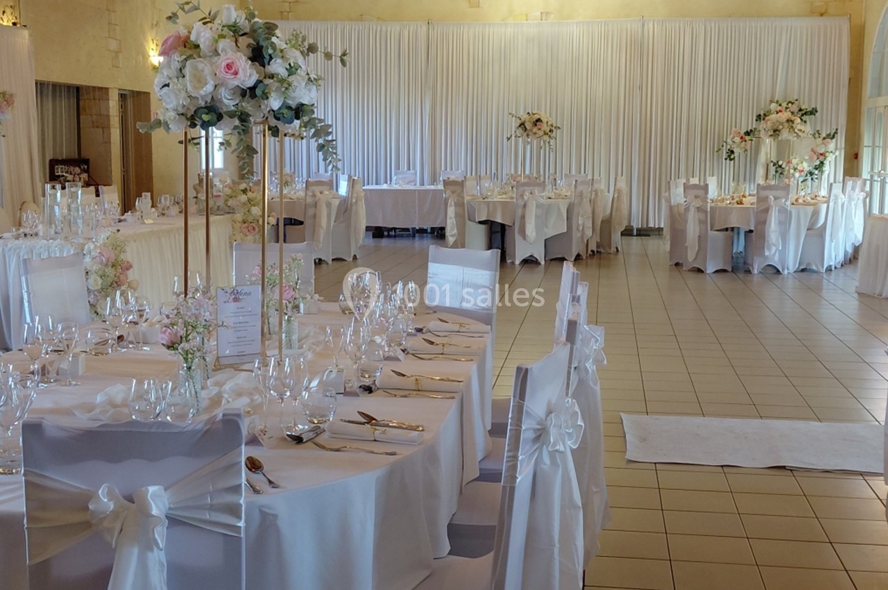 Salle de réception décorée pour un mariage, avec tables rondes dressées, nappes blanches et compositions florales.