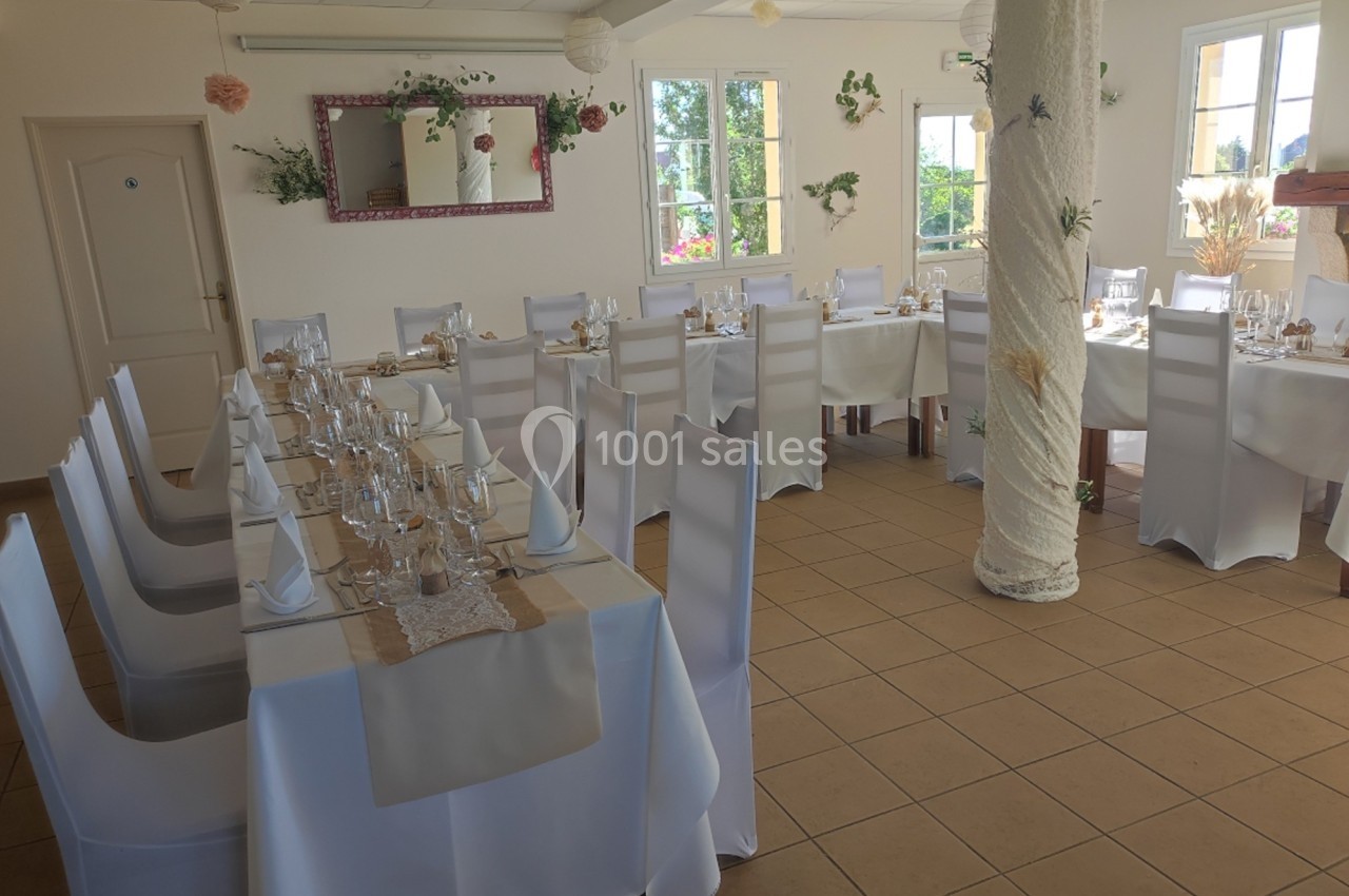 Salle lumineuse décorée pour un repas, avec des tables alignées, nappes blanches et chaises recouvertes de housses.