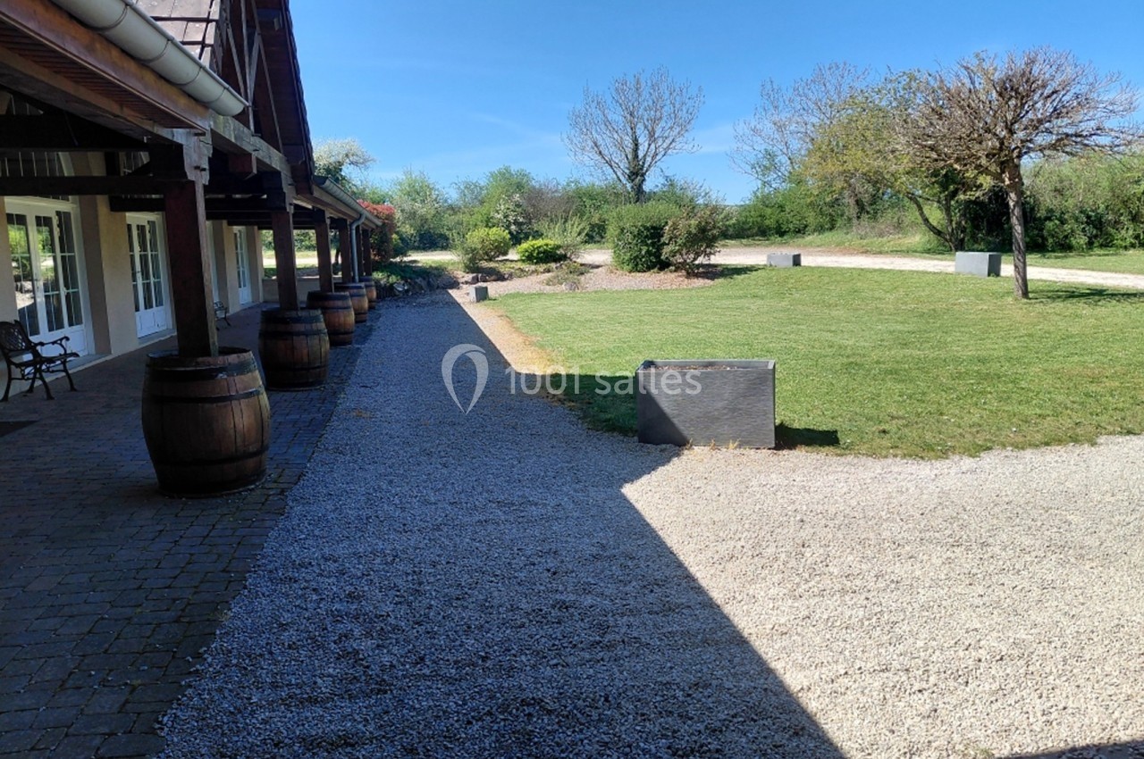 Allée en gravier bordée de tonneaux et pelouse, longeant un bâtiment avec des fenêtres sous un ciel dégagé.