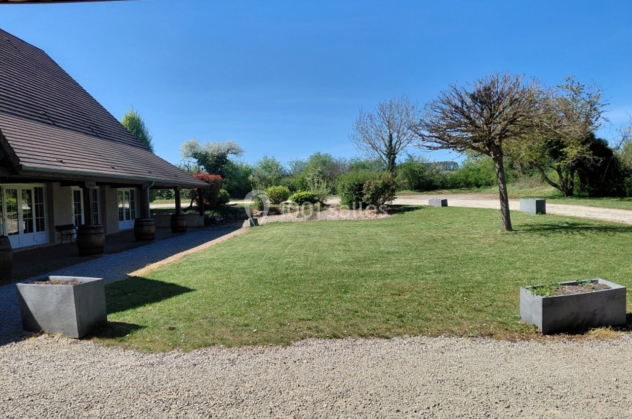 Vue d'une maison avec un toit en pente, entourée d'un jardin arboré et de bacs à fleurs sur une allée gravillonnée.