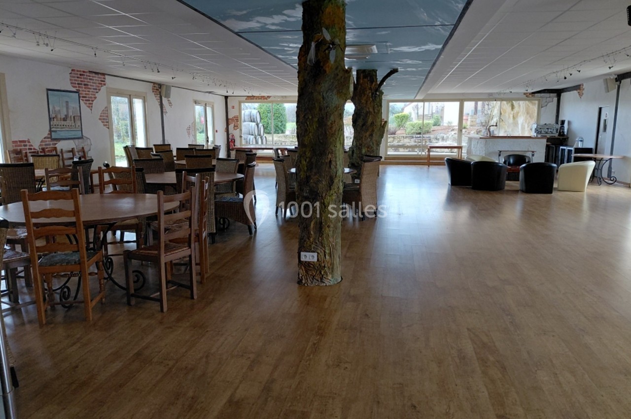 Salle spacieuse avec parquet, tables et chaises en bois, piliers décorés et grandes fenêtres donnant sur l'extérieur.