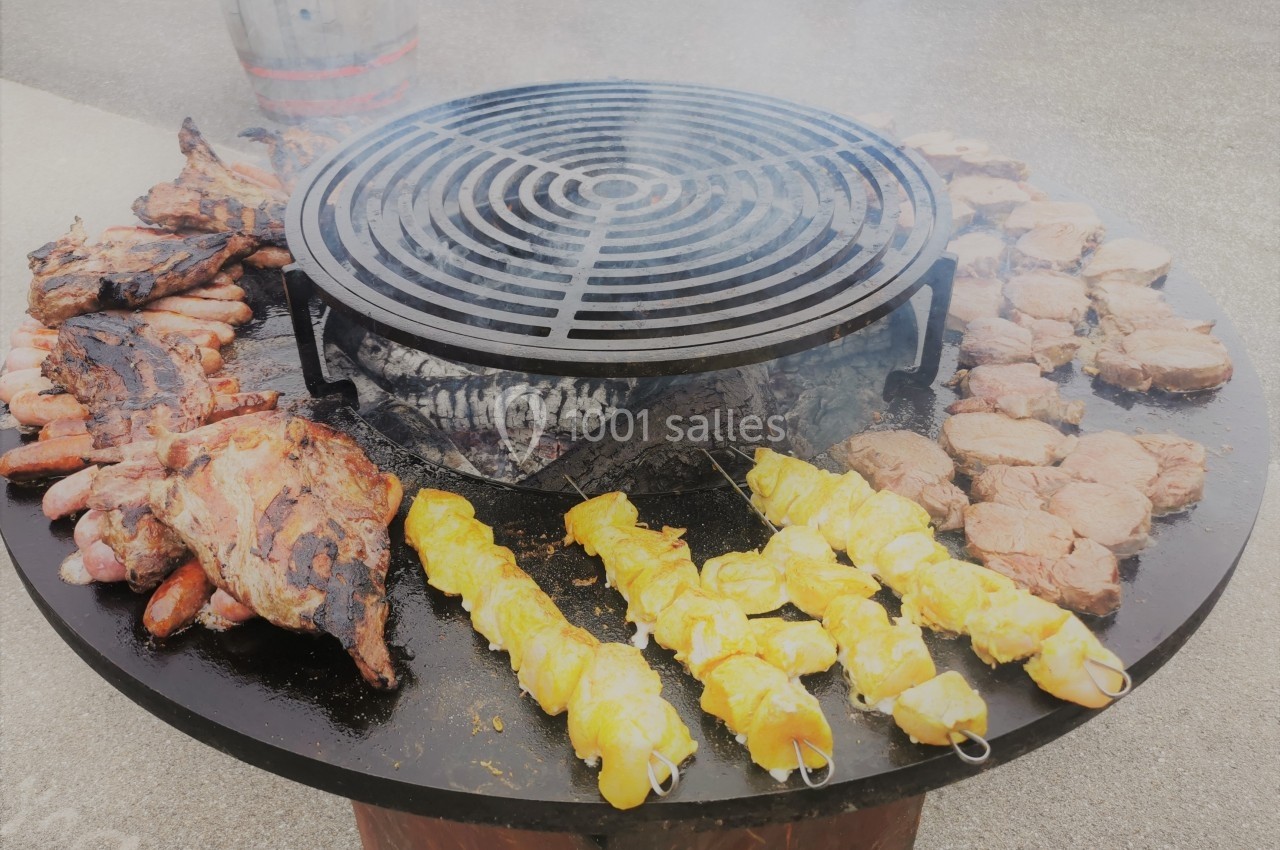 Grill circulaire avec diverses viandes en cuisson, dont brochettes de poulet, saucisses et steaks.