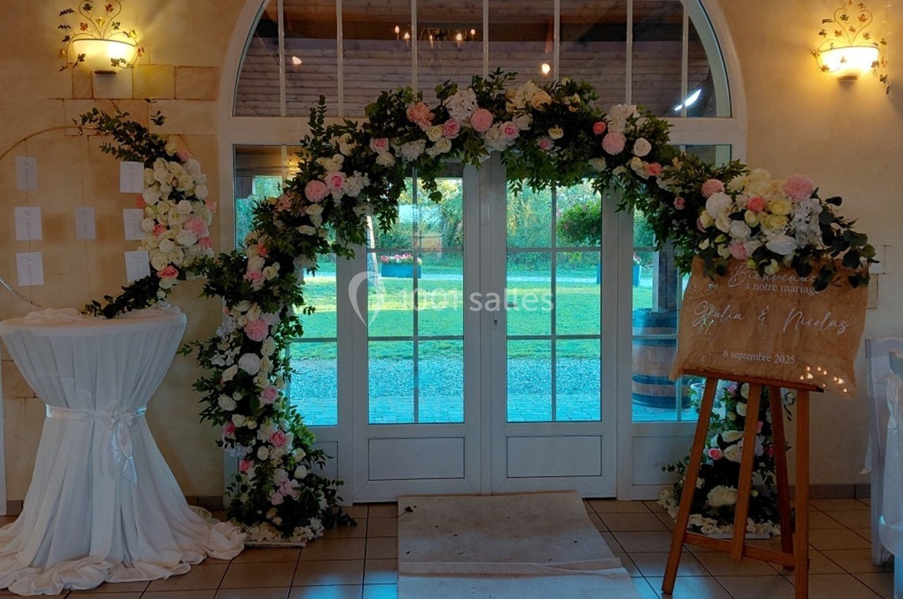 Arche florale décorative devant une porte vitrée, avec un panneau en bois mentionnant un événement et une table drapée.