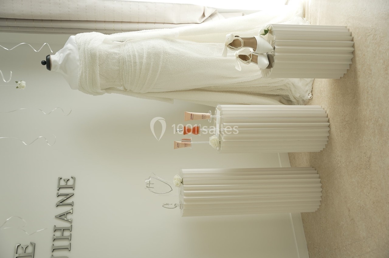 Mannequin portant une robe blanche, entouré de colonnes décoratives avec chaussures et produits cosmétiques exposés.