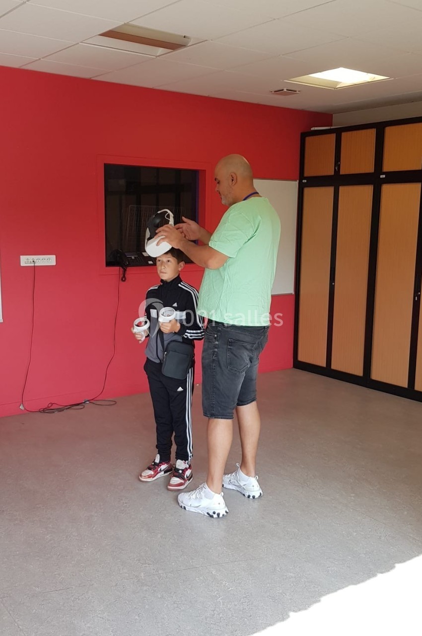 Un homme aide un enfant à ajuster un casque de réalité virtuelle dans une pièce aux murs rouges.