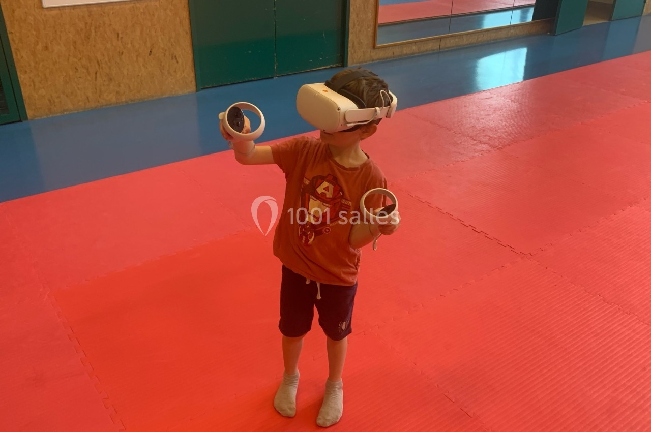 Un enfant portant un casque de réalité virtuelle et tenant deux manettes, debout sur un tapis rouge dans une salle.