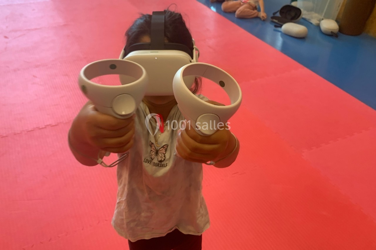 Enfant portant un casque de réalité virtuelle et tenant deux manettes sur un tapis de sol rose.