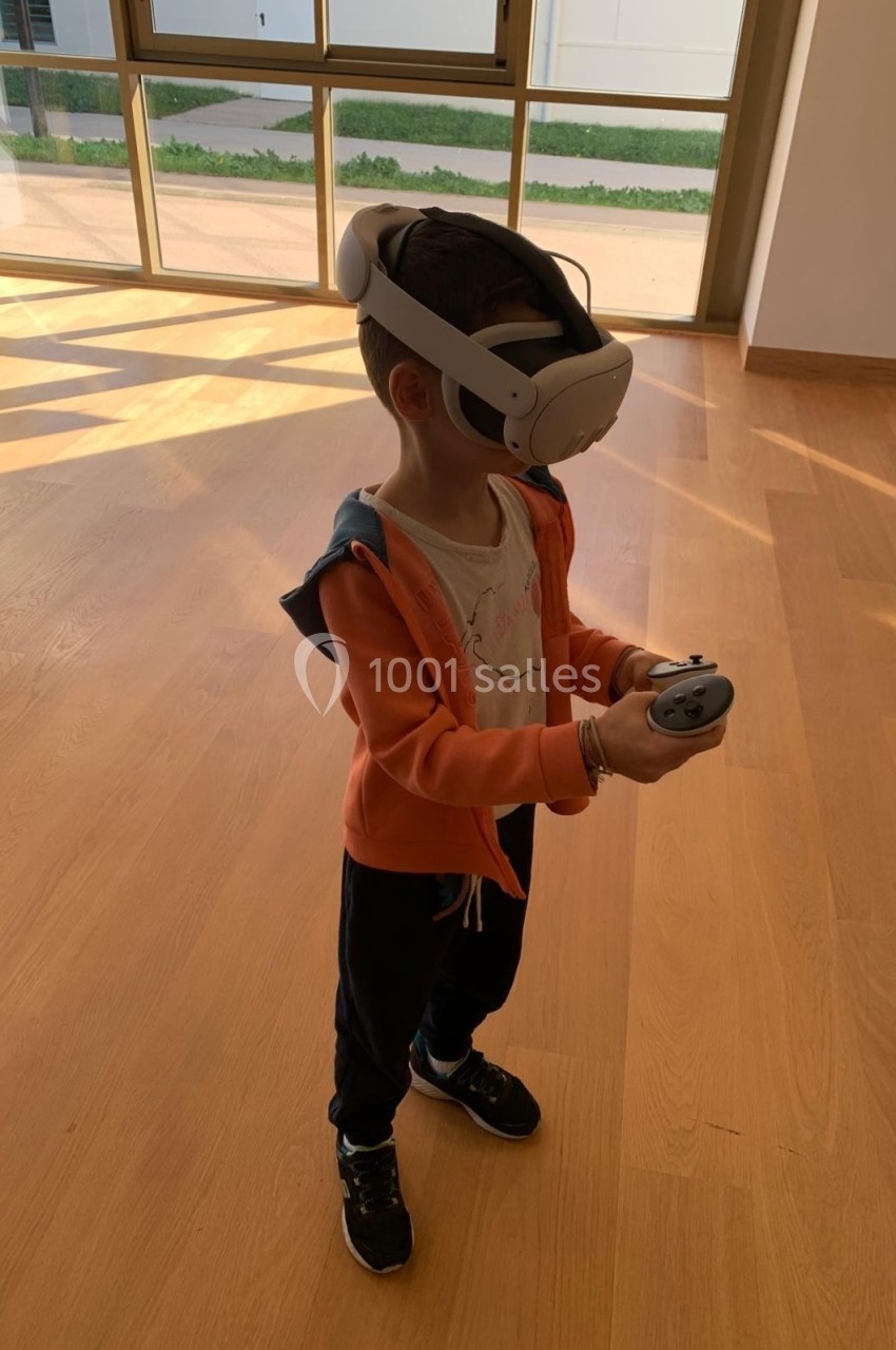 Un enfant portant un casque de réalité virtuelle et tenant une manette dans une pièce lumineuse au sol en bois.