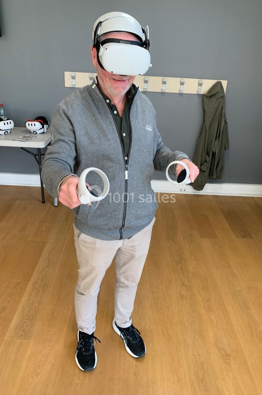 Homme portant un casque de réalité virtuelle et tenant deux manettes dans une pièce au sol en bois.