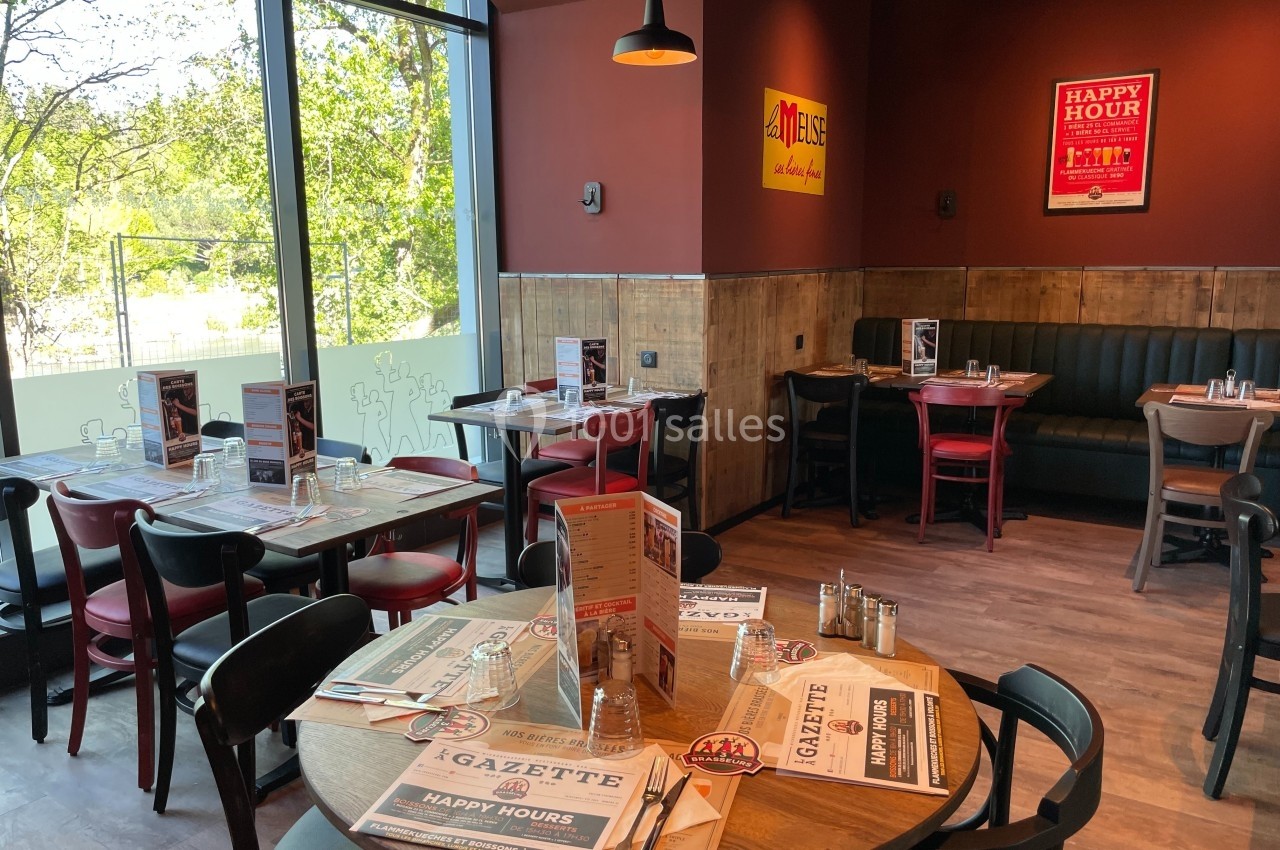 Salle de restaurant lumineuse avec tables dressées, chaises en bois et banquettes, décorée de cadres et affiches au mur.