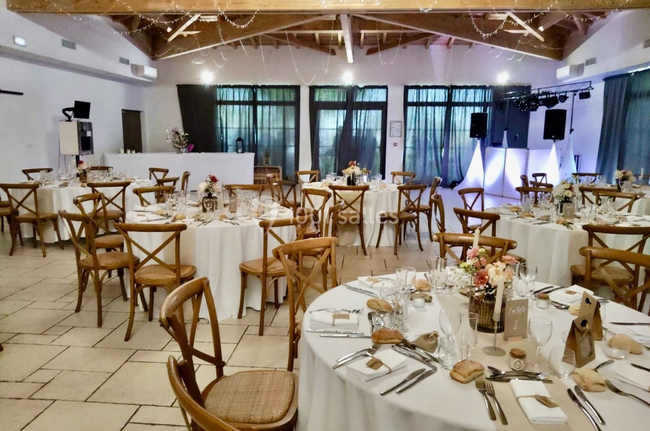 Salle de réception décorée pour un événement, avec tables rondes dressées, chaises en bois et éclairage tamisé.