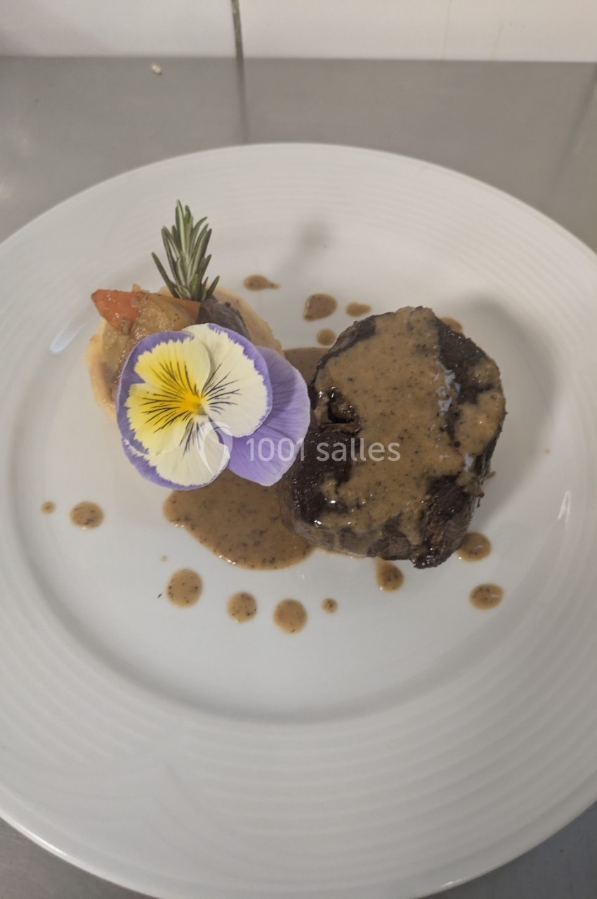 Assiette contenant un morceau de viande nappé de sauce, accompagné de purée, légumes et décorée d'une fleur comestible.