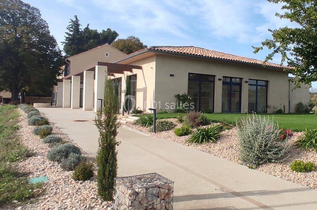 Bâtiment moderne avec façade beige, grandes baies vitrées, allée pavée et jardin paysager environnant.
