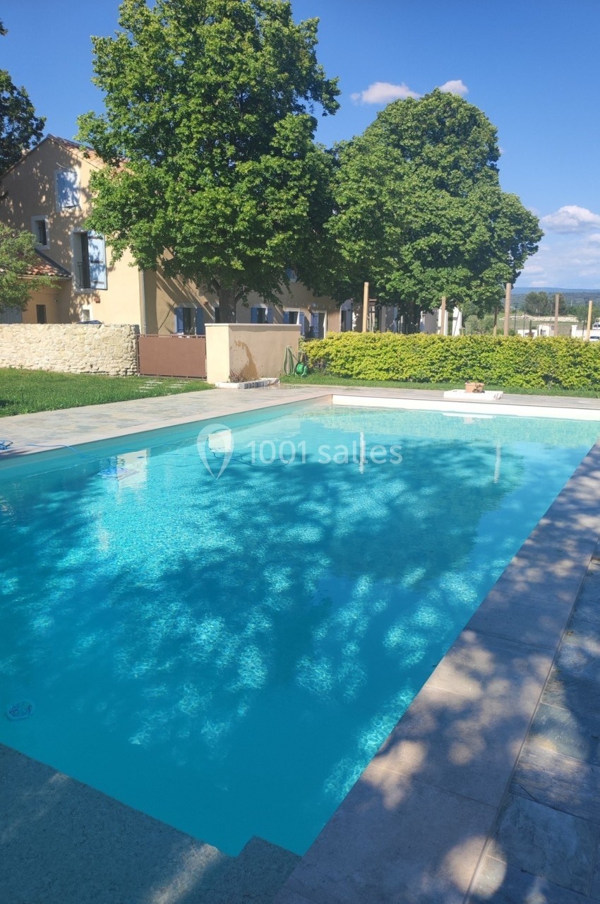 Piscine extérieure entourée de verdure et de bâtiments, sous un ciel bleu dégagé.