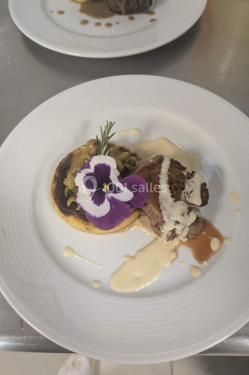 Assiette gastronomique avec viande, purée, sauce, légumes et décoration florale sur une table en métal.