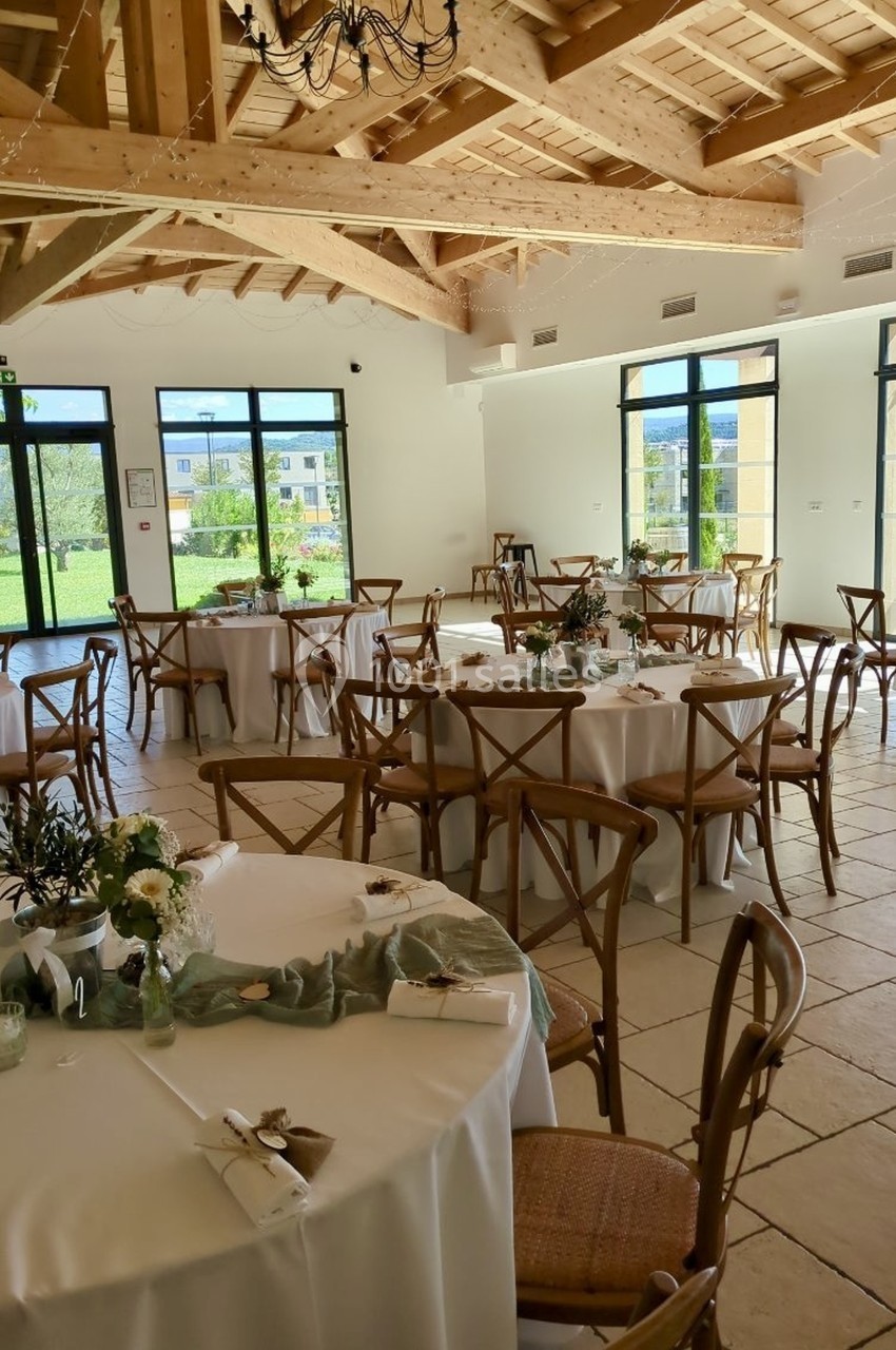 Salle lumineuse avec tables rondes décorées pour un événement, chaises en bois et vue sur un jardin à travers de grandes…