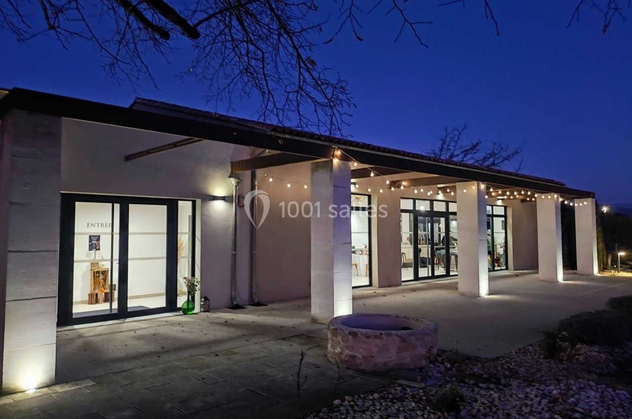 Façade d'un bâtiment moderne éclairé avec des guirlandes lumineuses, entouré de végétation et d'un puits en pierre.