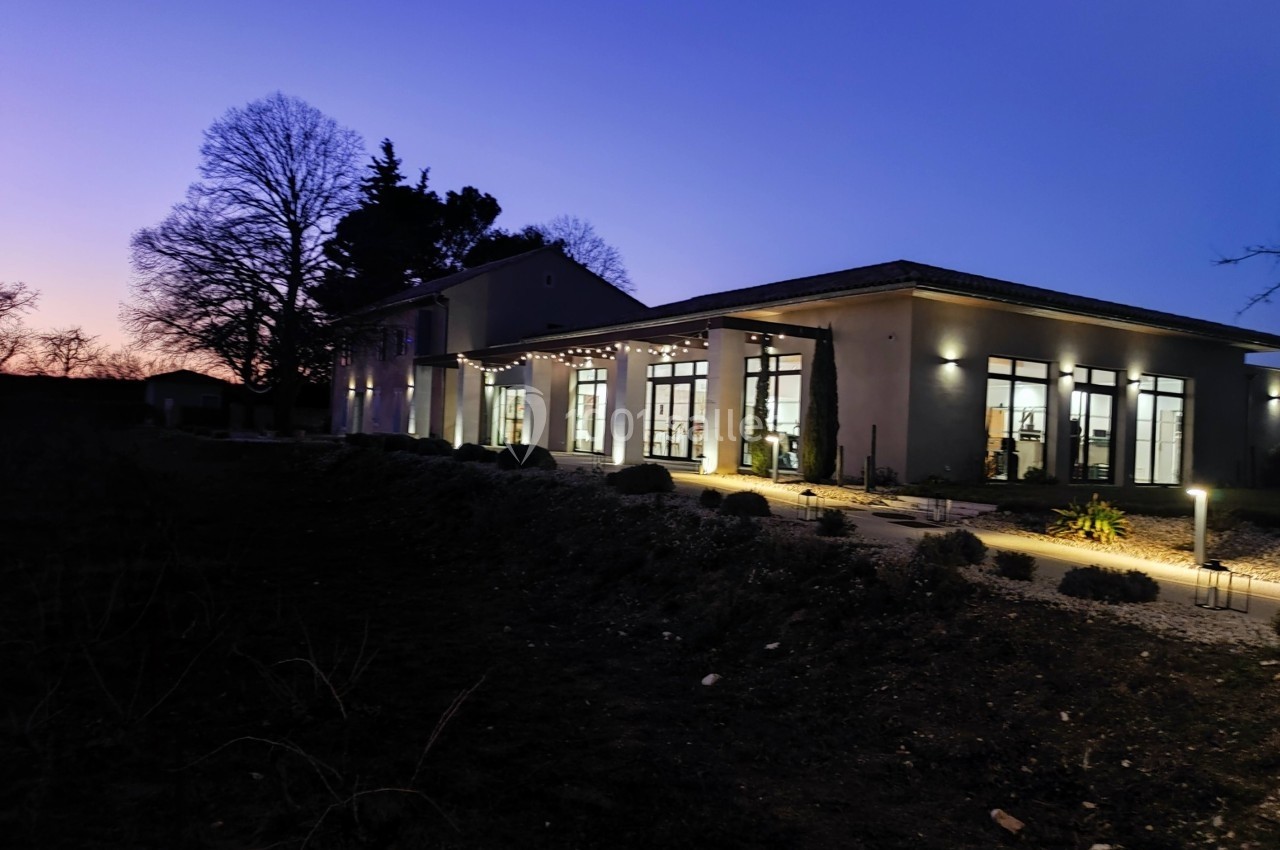 Bâtiment moderne éclairé en soirée, entouré d'arbres et de végétation sous un ciel crépusculaire.