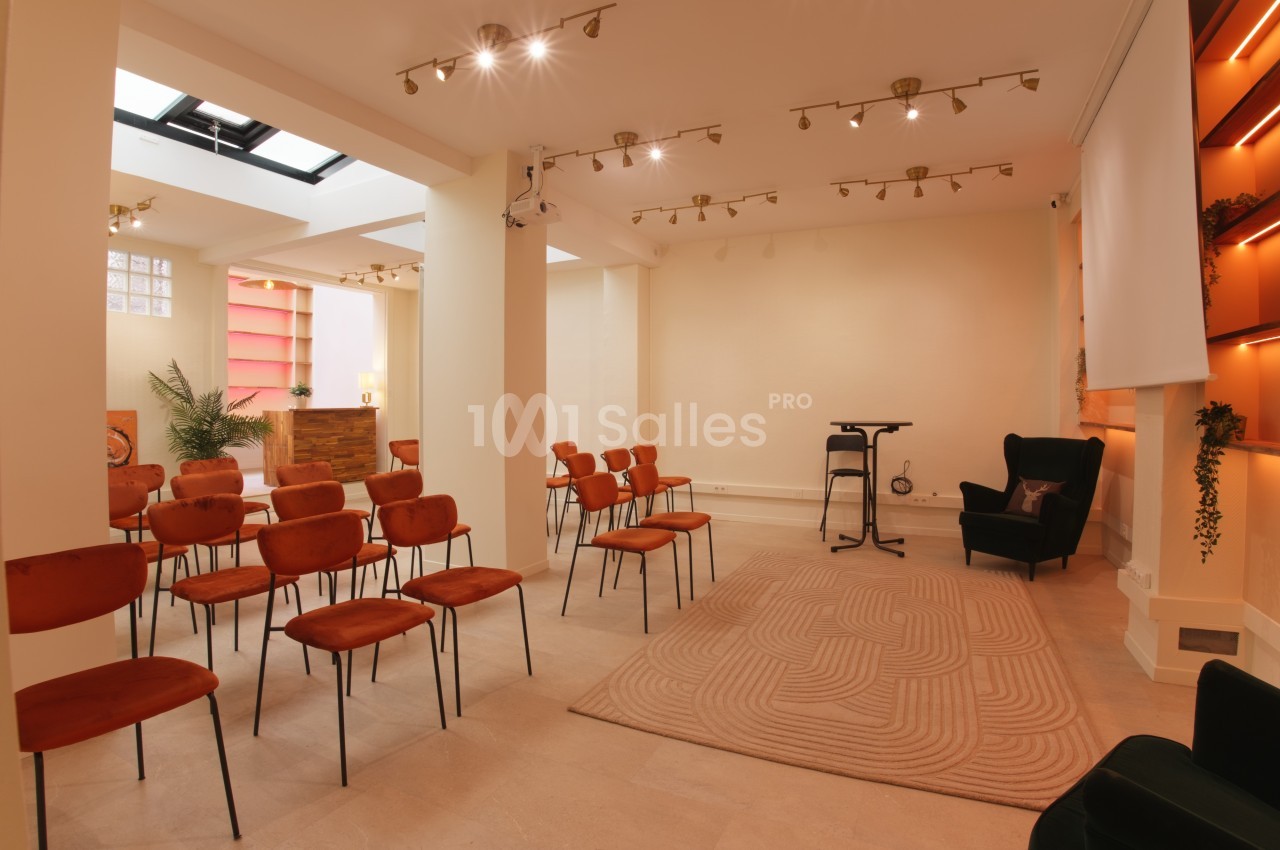 Salle lumineuse avec des chaises orange alignées, un tapis beige, un fauteuil noir et un bar en arrière-plan.