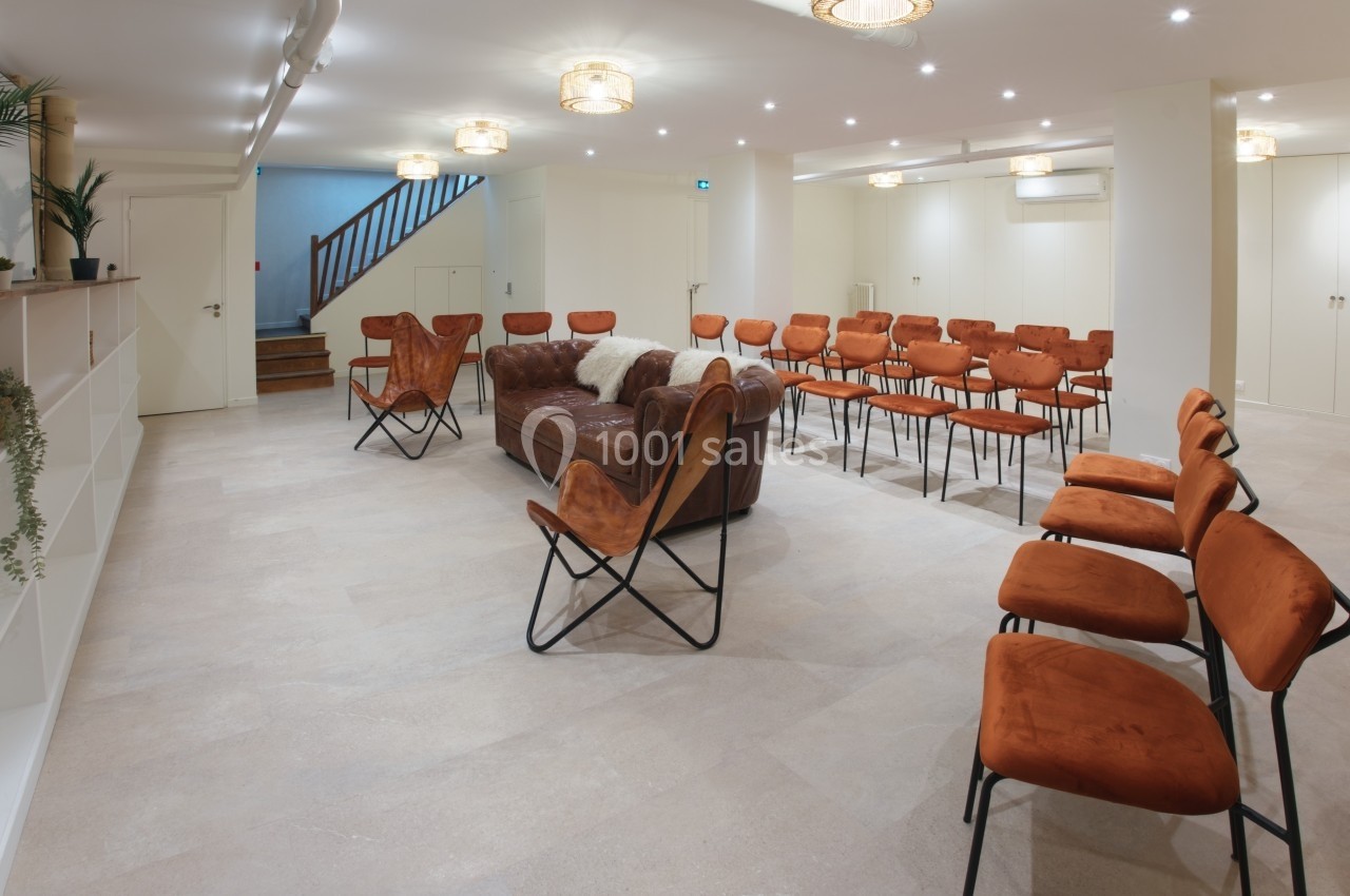 Salle lumineuse avec des chaises orange disposées en rangées, un canapé en cuir et un escalier en arrière-plan.