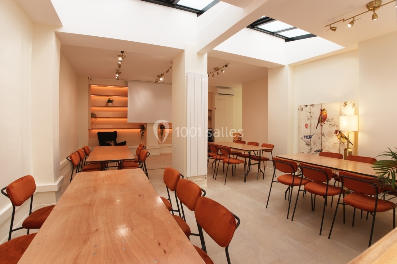 Salle lumineuse avec tables en bois, chaises orange, étagères éclairées et plafond avec puits de lumière.