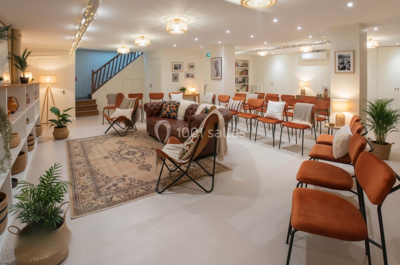 Salle lumineuse aménagée avec des fauteuils orange, un canapé, des plantes et un tapis, idéale pour des réunions.