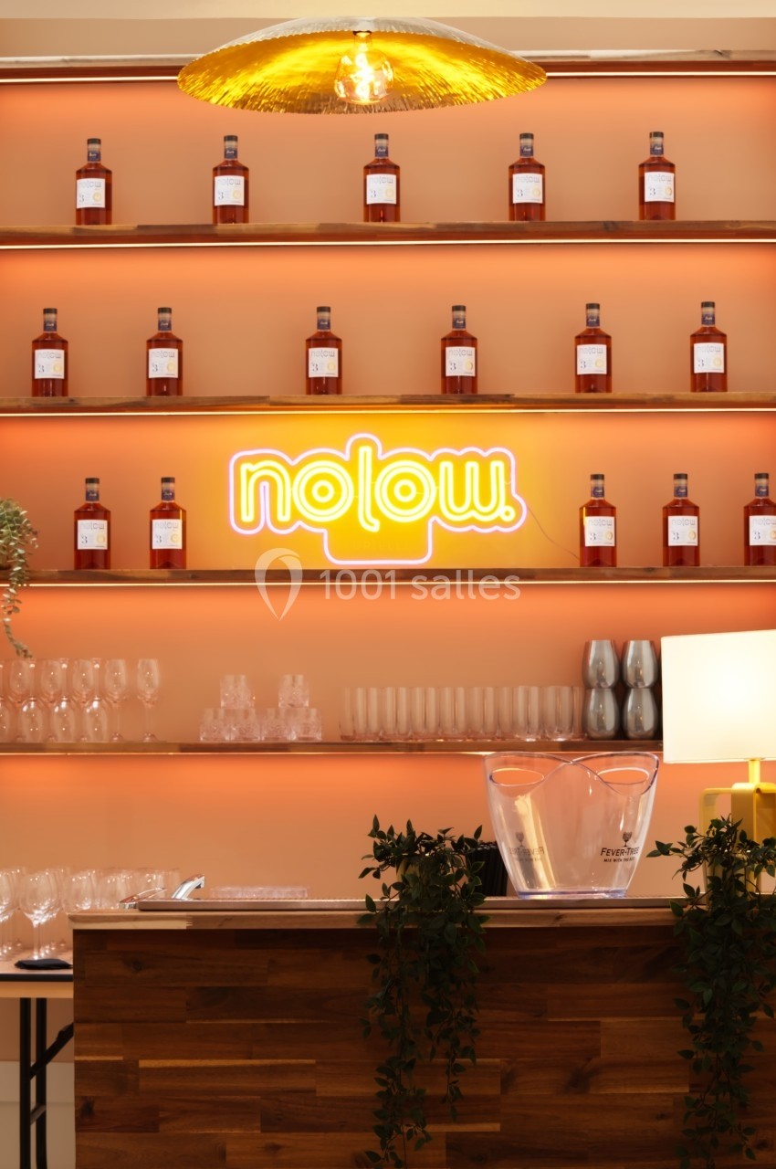 Bar en bois avec étagères illuminées exposant des bouteilles, un néon ’nolow’ et des verres disposés en dessous.