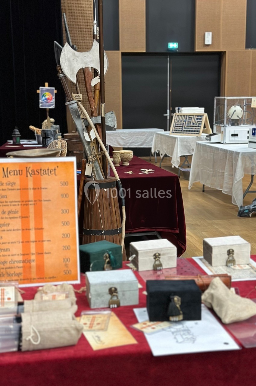 Stand avec jeux en bois, énigmes et accessoires médiévaux, accompagné d'une liste de tarifs sur une affiche orange.