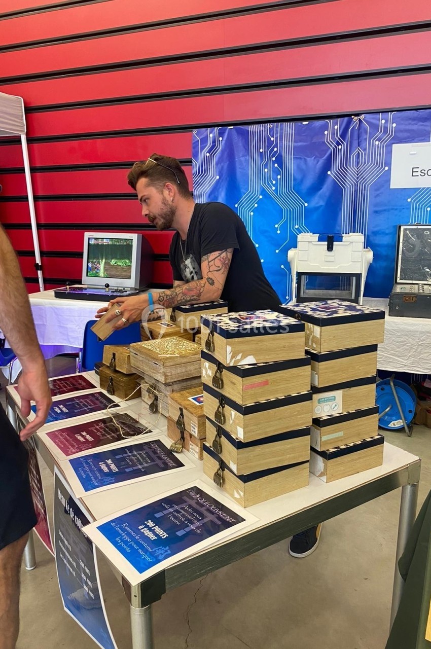 Un homme présente des boîtes en bois et des affiches sur une table lors d'un événement dans un espace intérieur.